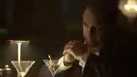 Szene aus der Serie "The Penguin": Oswald "Oz" Cobblepot (Colin Farrell) sitzt, während er einen Martini trinkt, mit übereinander liegenden Händen und einem ernsten Blick in einer Bar.