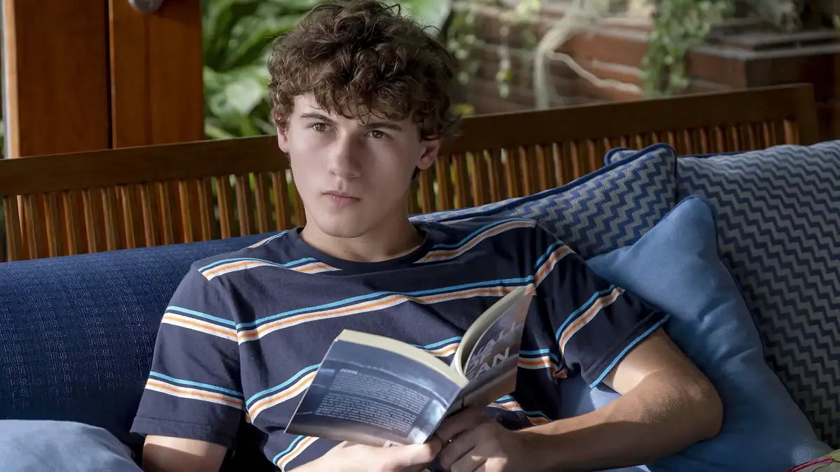 Szene aus der HBO-Serie The White Lotus: Ein junge Mann in einem gestreiften Shirt sitzt mit einem Buch auf dem Sofa der Terrasse und schaut kritisch in die Ferne .