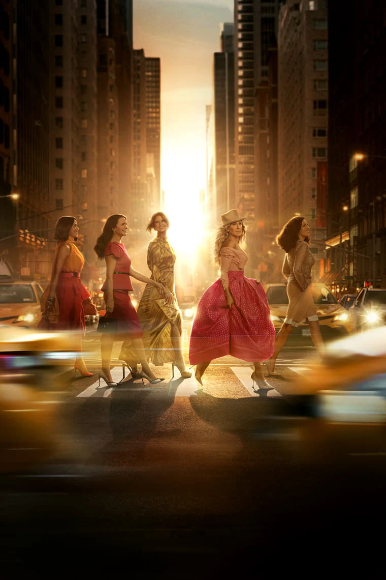 5 women from the cast in colourful dresses crossing a busy street with yellow taxis and tall buildings in the background